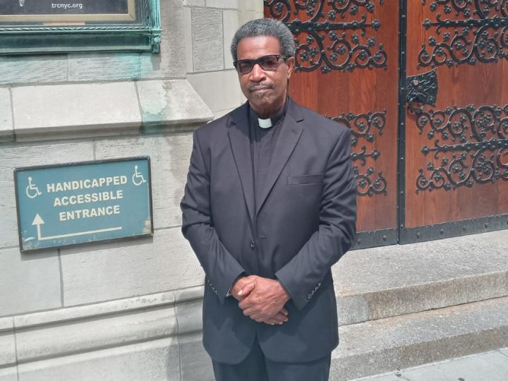 A middle-aged Black man with brown skin, salt-and-pepper curly hair stands in front of church doors. He is dressed in a black pastor's outfit and black shoes. He is holding his hands together in front of him.