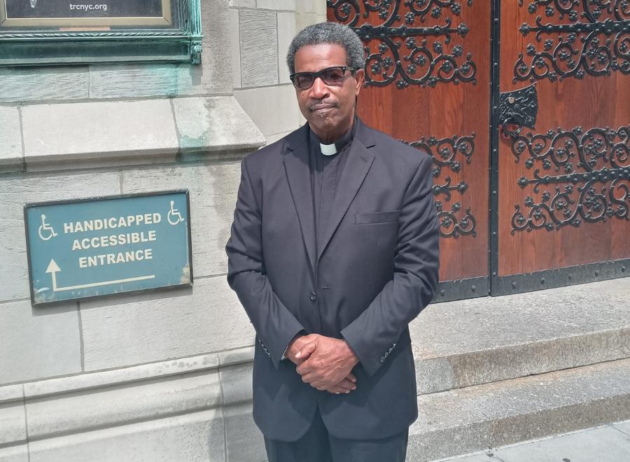 A middle-aged Black man with brown skin, salt-and-pepper curly hair stands in front of church doors. He is dressed in a black pastor's outfit and black shoes. He is holding his hands together in front of him.