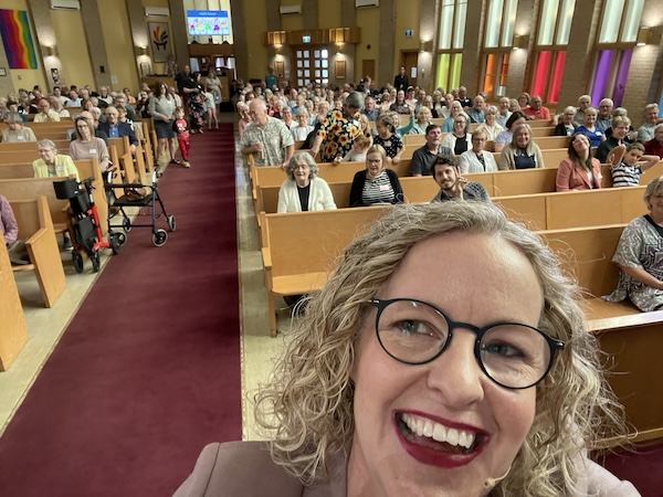 A white woman with black-framed glasses, curly blonde hair, red lipstick smiles in a selfie in church. Behind her are pews and people sitting and smiling