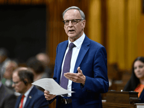 A middle-aged white man speaks in Parliament. He has white hair, black-framed glasses, is wearing a blue suit and purple tie. He is holding a paper in his right hand and gesturing with his left hand