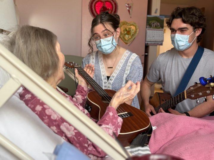 A senior woman lies in a hospital bed, holding a ukulele and speaking with two young musicians who are wearing medical masks. One of the musicians also holds a ukulele, while the other has a guitar. They are engaged in warm conversation.
