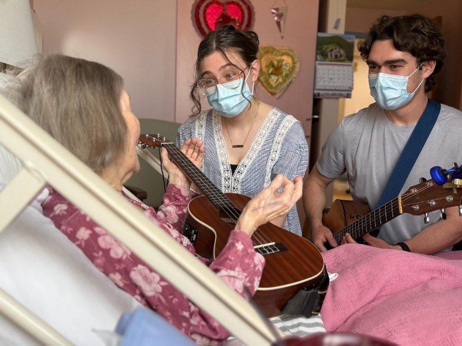 A senior woman lies in a hospital bed, holding a ukulele and speaking with two young musicians who are wearing medical masks. One of the musicians also holds a ukulele, while the other has a guitar. They are engaged in warm conversation.