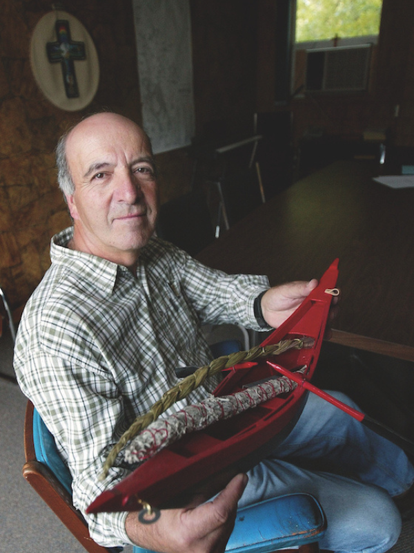 A middle-aged Indigenous man sits in a. chair. He is balding, wearing a plaid shirt and holding a canoe in his hands