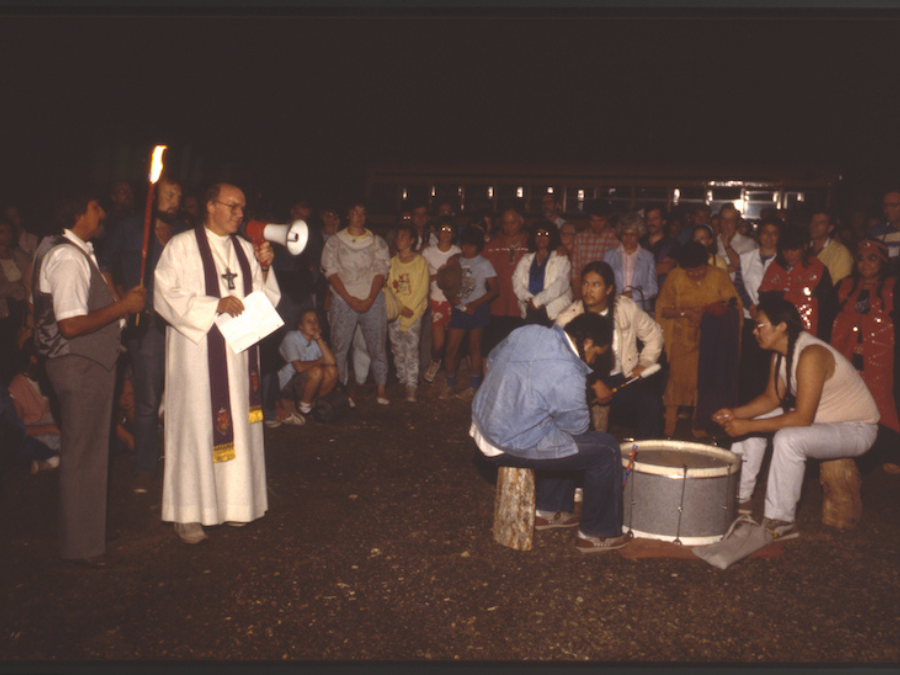A white priest in white robes and a cross holds a megaphone in his left hand and a paper in his right hand. A group of Indigenous folks are standing behind him. Two are sitting to his left, around a drum