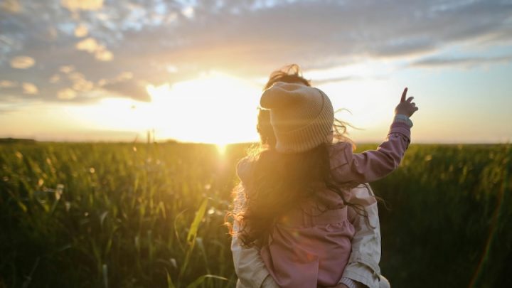 A person holds a child against a sunset. Their backs are facing the camera. The child has her right arm up and is pointing at the sky.