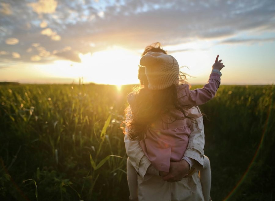 A person holds a child against a sunset. Their backs are facing the camera. The child has her right arm up and is pointing at the sky.