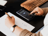 A white woman's hands with the right on a black calculator and the left is wearing a watch, holding a pen writing on a piece of paper