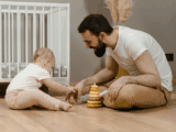 A white man with dark brown hair and beard sits on the floor playing with a baby. They are both dressed in white t-shirts and tan pants