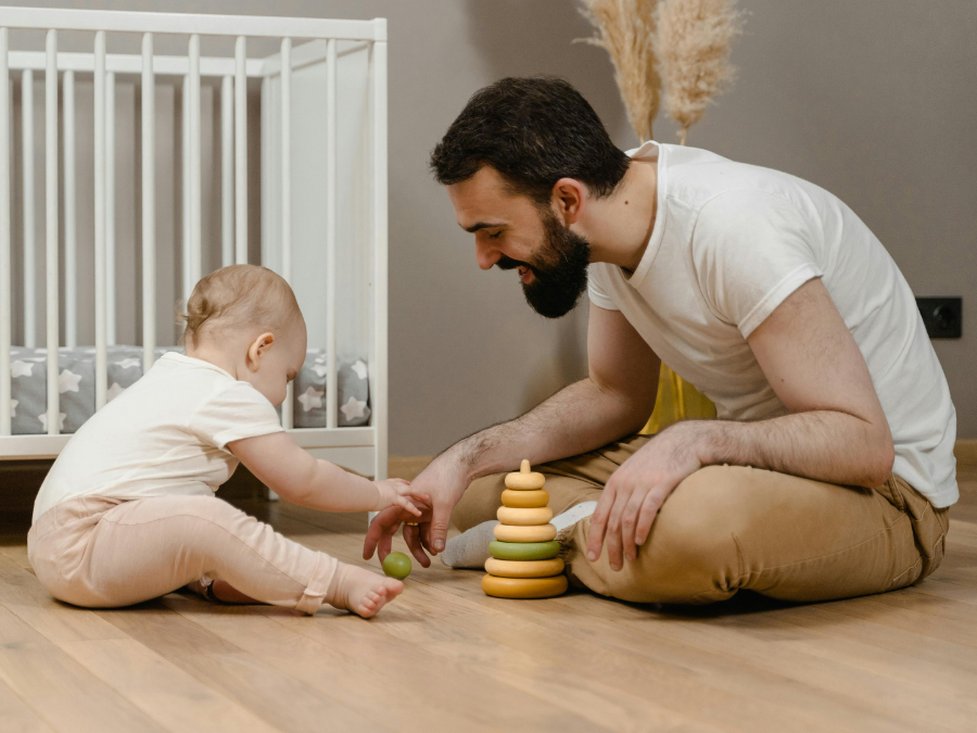 A white man with dark brown hair and beard sits on the floor playing with a baby. They are both dressed in white t-shirts and tan pants