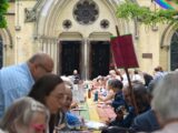 Dozens of people sit at a long, rainbow-painted picnic table outside Metropolitan United Church in Toronto, sharing food and conversation during a community potluck.