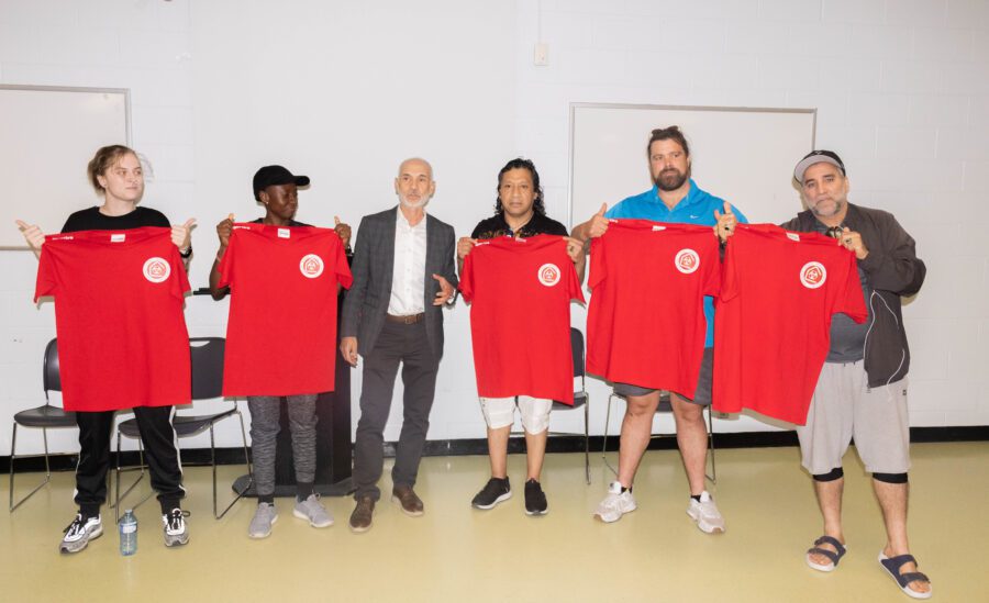 Six people stand in a row indoors, holding up red Canadian Street Soccer Association jerseys. Hossam Khedr, wearing a suit, stands in the centre.