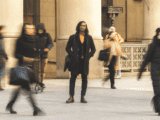 A Black man with long black hair stands in front of a building. The people walking behind him are blurry. He is wearing a black jacket, dark jeans, brown boots and has a black bag