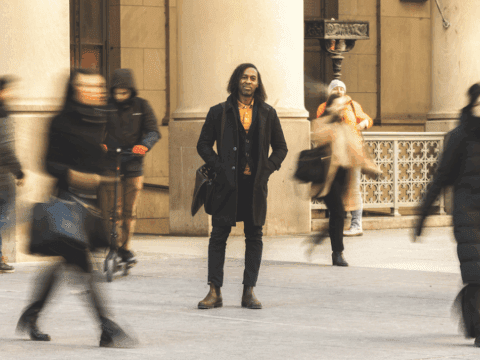 A Black man with long black hair stands in front of a building. The people walking behind him are blurry. He is wearing a black jacket, dark jeans, brown boots and has a black bag