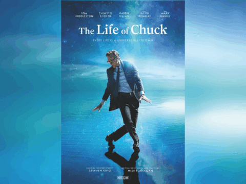 A white man wearing glasses, in a blue suit, is dancing against a background of blue ocean and blue sky