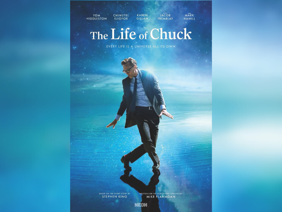 A white man wearing glasses, in a blue suit, is dancing against a background of blue ocean and blue sky