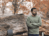 A brown-skinned man with dark brown hair, facial hair and glasses poses by a tree trunk. He is wearing an olive-coloured shirt and has his arms crossed in front of him