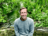 Headshot of a white man with brown hair in a grey sweater and khaki pants. He is sitting on a log and behind him is a river and green bush