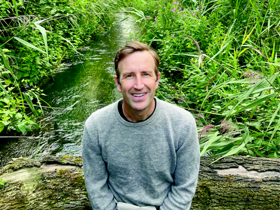 Headshot of a white man with brown hair in a grey sweater and khaki pants. He is sitting on a log and behind him is a river and green bush