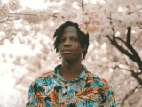 Headshot of a Black man with dreads. He is wearing a colourful button up shirt with palm leaves. He is standing against a backdrop of cherry blossoms