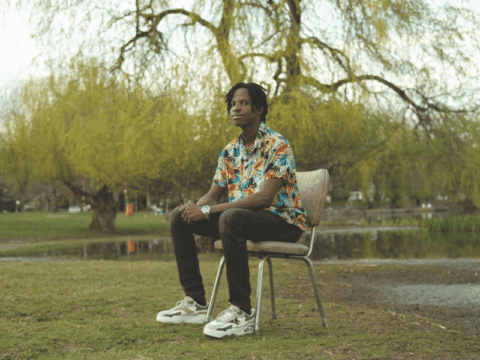 A Black man with dreadlocks sits on a chair in front of cherry blossom trees. He is wearing a colourful button-up shirt, a watch on his left hand, black jeans and white sneakers.