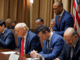 A group of white men sit with heads bowed. One of them has white hair, a red tie and blue suit. Next to him is a man with brown hair, blue suit and tie. His hands are clasped. Behind them stands a Black man in a blue suit and red tie, head bowed.