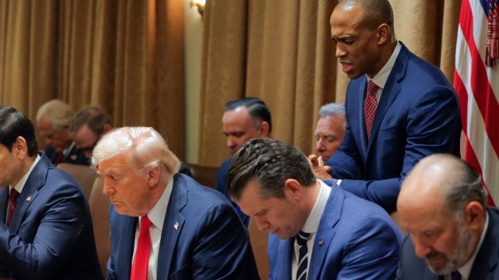 A group of white men sit with heads bowed. One of them has white hair, a red tie and blue suit. Next to him is a man with brown hair, blue suit and tie. His hands are clasped. Behind them stands a Black man in a blue suit and red tie, head bowed.