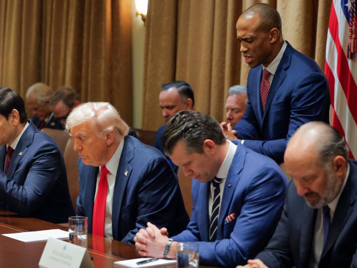 A group of white men sit with heads bowed. One of them has white hair, a red tie and blue suit. Next to him is a man with brown hair, blue suit and tie. His hands are clasped. Behind them stands a Black man in a blue suit and red tie, head bowed.