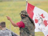 A masked Indigenous protester wearing camouflage clothing and a hat gestures while standing beside a young boy. Behind them, a flag featuring a red maple leaf and an image of a First Nations person in traditional regalia flutters in the wind. The background shows a blurred green field.