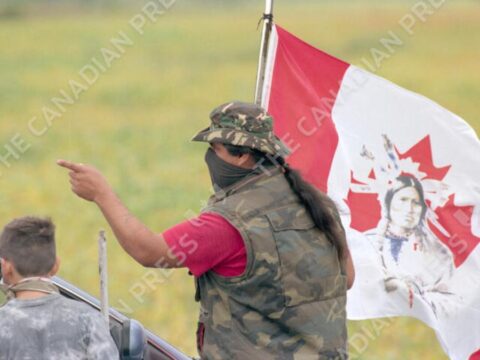 A masked Indigenous protester wearing camouflage clothing and a hat gestures while standing beside a young boy. Behind them, a flag featuring a red maple leaf and an image of a First Nations person in traditional regalia flutters in the wind. The background shows a blurred green field.