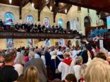 People stand in a church with stained-glass windows. Some wave ribbons
