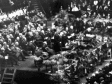 A black-and-white photo shows people sitting in bleachers and in chairs on a stage as other people in church robes stand.