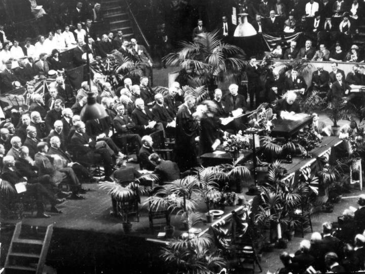 A black-and-white photo shows people sitting in bleachers and in chairs on a stage as other people in church robes stand.