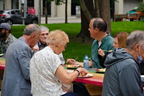 A group of people, including older and younger adults, sit at a long picnic table in front of Metropolitan United Church in Toronto, sharing a meal and conversing. The outdoor setting features green grass, trees, and nearby buildings in the background