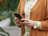 Close up of a white woman's hands. She is wearing a floral dress and an orange cardigan. On her right hand is a gold watch. She is holding a black phone in her left hand