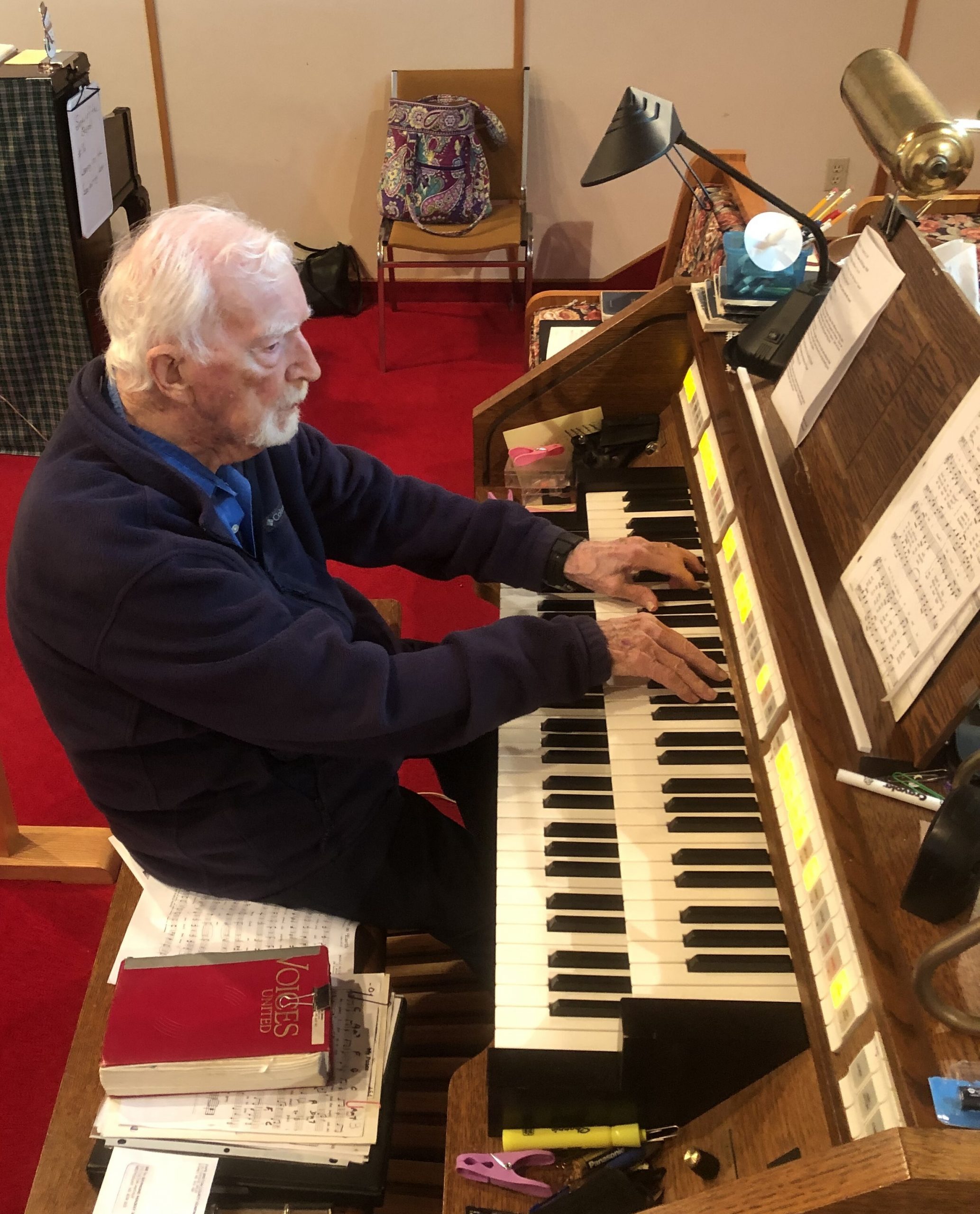 Ray MacKay, 102, plays the organ at Westmount United Church in Nova Scotia, seated at the console with sheet music and a red Voices United hymnal nearby.