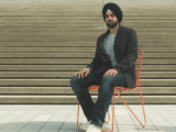 A young brown-skinned man in a turban sits on a chair in front of steps. He is wearing a black turban, has a short beard, in a light grey shirt, brown blazer and jeans. He has white Chucks on