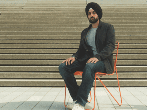 A young brown-skinned man in a turban sits on a chair in front of steps. He is wearing a black turban, has a short beard, in a light grey shirt, brown blazer and jeans. He has white Chucks on