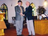 Two middle-aged white men shake hands. They are standing in a church, behind them are stained glass windows, a pulpit. One man is in grey slacks and a grey blazer. The other man is in light grey slacks and a black blazer.