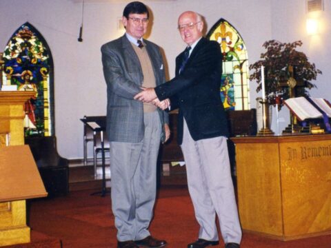 Two middle-aged white men shake hands. They are standing in a church, behind them are stained glass windows, a pulpit. One man is in grey slacks and a grey blazer. The other man is in light grey slacks and a black blazer.