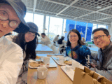 Four East Asian people gather around a table and smile. They have food and drinks in front of them. There are two men with glasses. One of them is wearing a blue hat. There are two women and one of them is wearing glasses. The other has long black hair and a blue hat on.