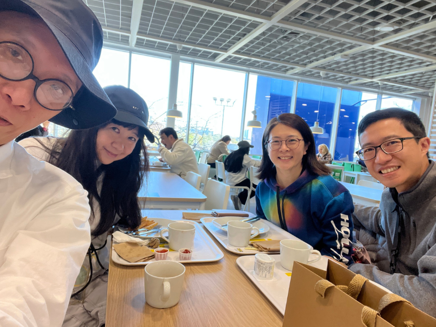Four East Asian people gather around a table and smile. They have food and drinks in front of them. There are two men with glasses. One of them is wearing a blue hat. There are two women and one of them is wearing glasses. The other has long black hair and a blue hat on.