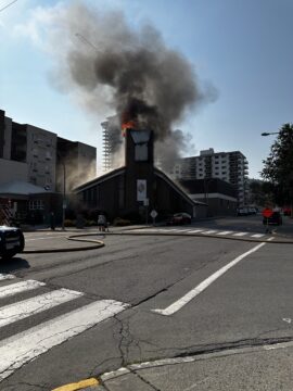 Kamloops United Church with flames and black smoke billowing from the roof, a fire hose stretched across the road, and nearby buildings visible under a clear sky.