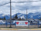 An empty lot with a fence around it. There is a sign that says, "It's gonna be okay" in black. The text has two red hearts around it. In the background are mountains and clouds.