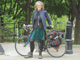 A white woman with curly grey hair and glasses stands before a bicycle. She is wearing a pastor's black top, a navy blazer, green skirt, black tights and black boots. She has glasses on and around her neck are a set of keys