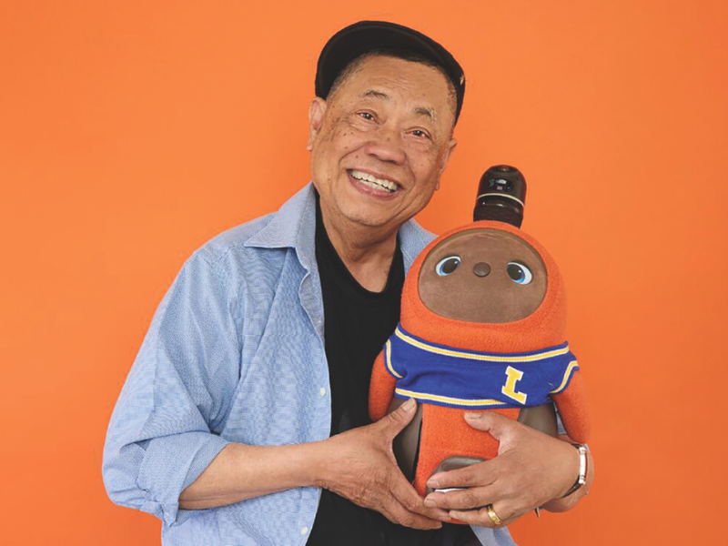 An older brown-skinned man with black hair holds a plushie robot in his arms. The man is wearing a black shirt with a blue button up. He is smiling. The robot has a brown face, orange limbs and wearing a blue shirt with an "L" on it.