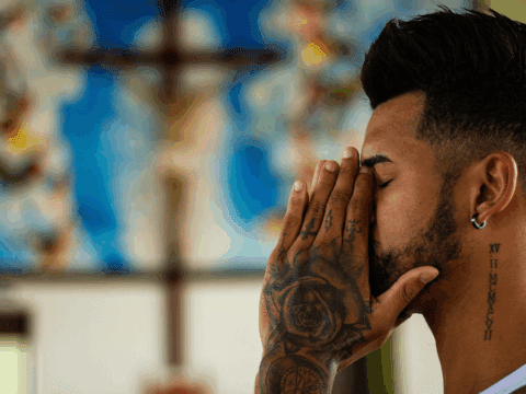 Side profile of a brown-skinned man with hand tattoos and a beard. His face is in his hands. In the background is a blurry crucifix