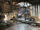 Interior of Kamloops United Church sanctuary severely damaged by fire, with charred walls, exposed beams, scattered debris, and standing water on the floor. A burned piano and broken furniture sit amid the wreckage.