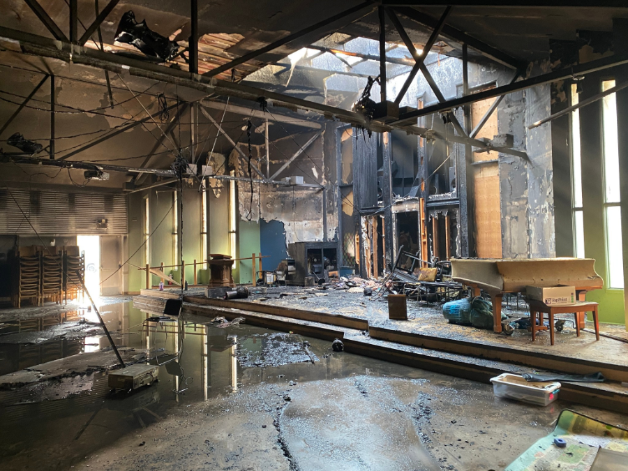 Interior of Kamloops United Church sanctuary severely damaged by fire, with charred walls, exposed beams, scattered debris, and standing water on the floor. A burned piano and broken furniture sit amid the wreckage.