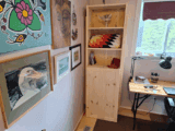 A wooden bookshelf made from a casket stands in the corner of a home office, surrounded by artwork and a desk by the window.