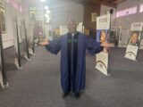 A Black man in ministers' robes stands inside a church, among a series of panels with photos on them